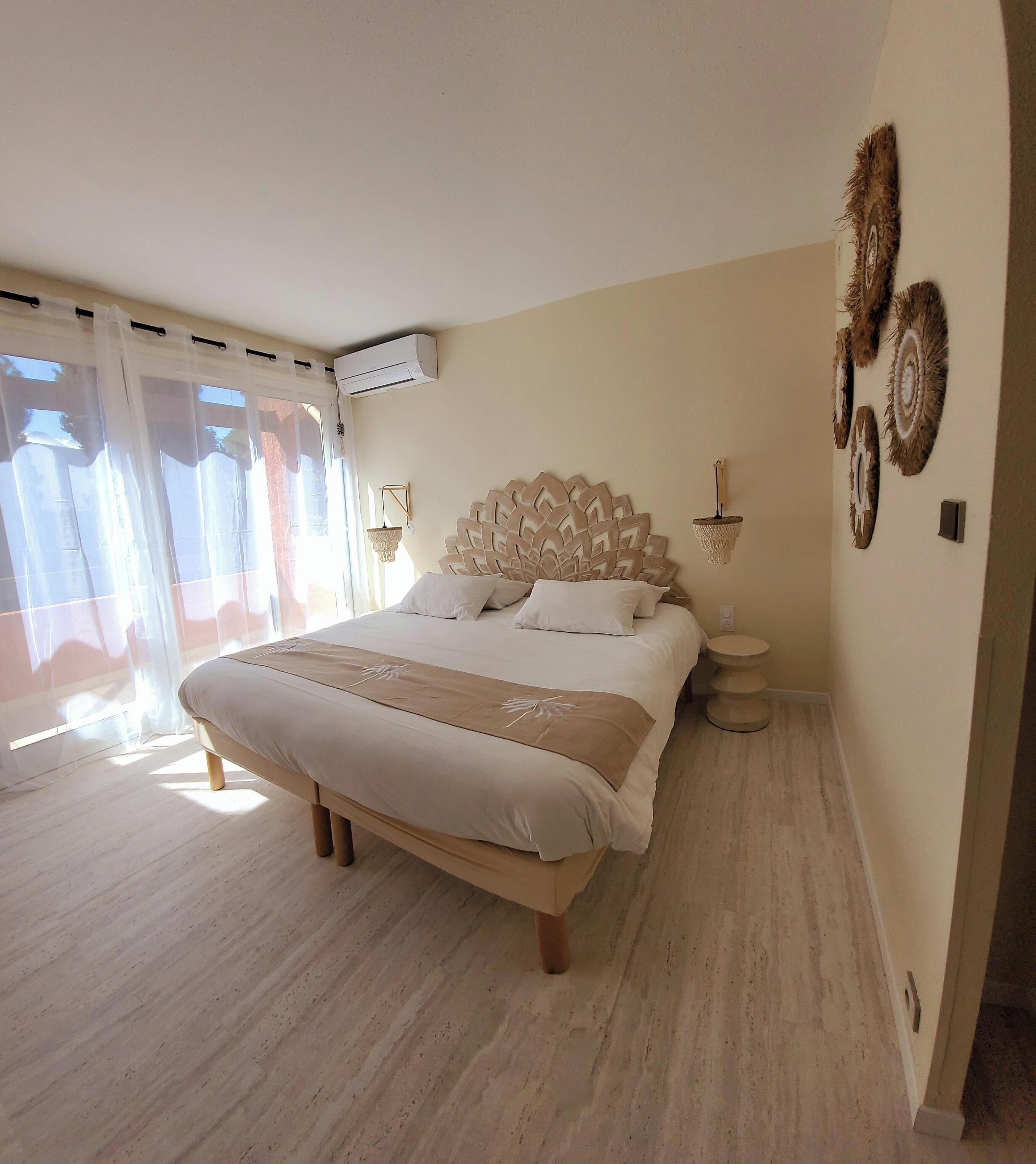 A bedroom with a large bed, a bedside table, a wall-mounted air conditioner, and a window with sheer curtains.