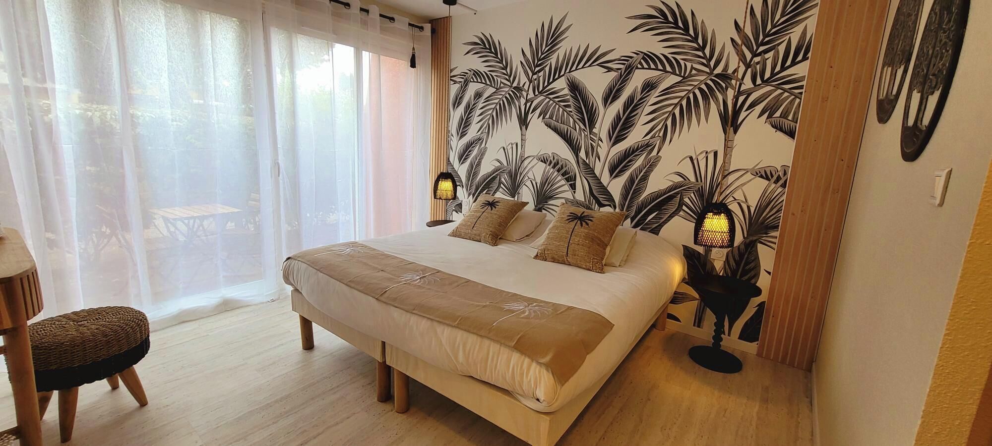 Un dormitorio con una cama, una silla, un ventanal con cortinas translúcidas y una pared con motivos de hojas tropicales.