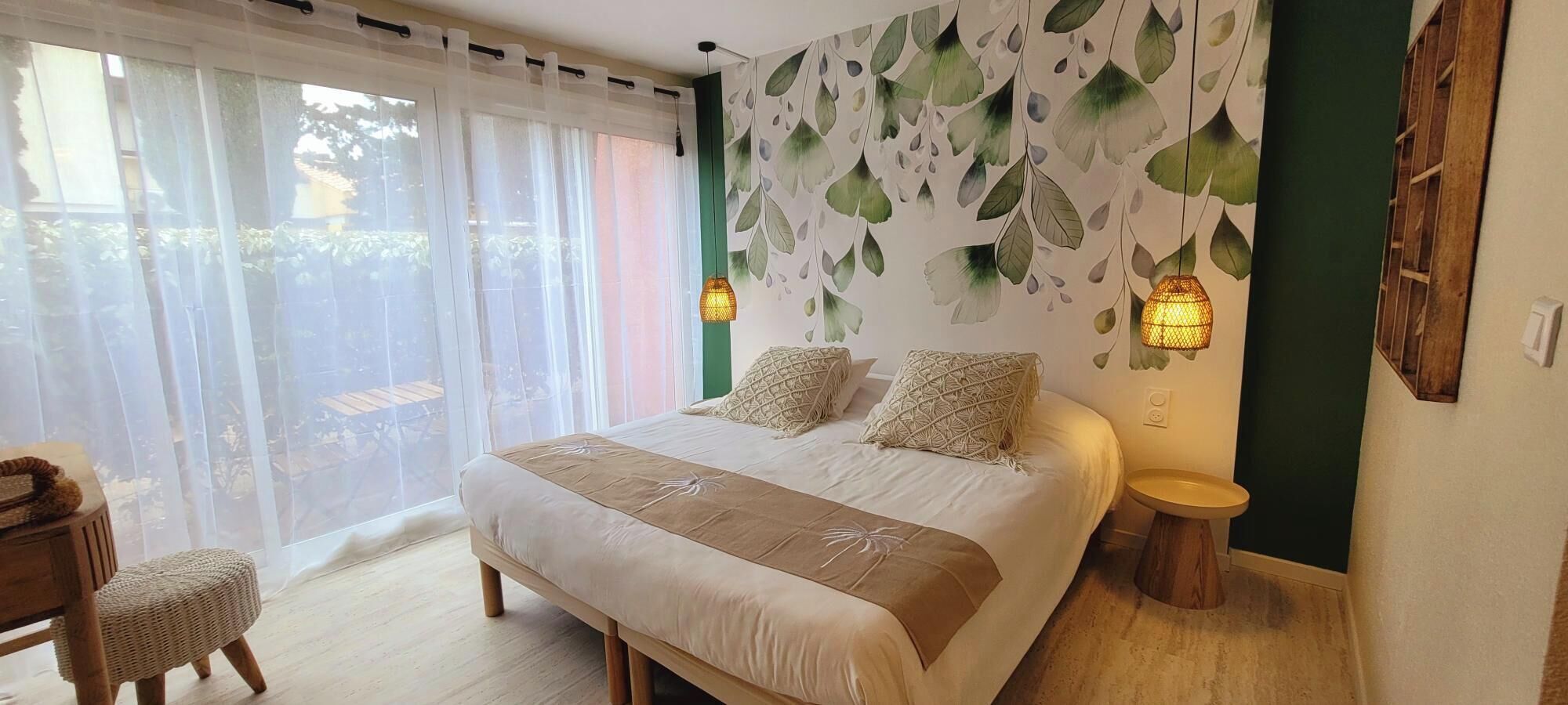 A bedroom with a large bed, two bedside lamps, a wooden side table, and a view of the outdoors through sheer curtains.
