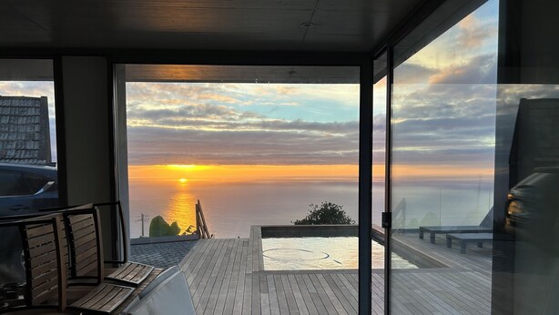 Un balcone con vista sul tramonto sul mare, arredi in legno e una porta vetrata che si apre sull'area esterna.