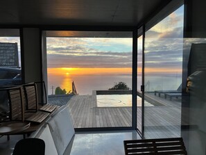 A balcony with a view of the sunset over the sea, wooden furniture, and a glass door leading to the outdoor area.