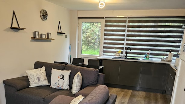 A modern living room with a grey sectional sofa, a small kitchen area with a sink and window blinds, and a wall-mounted clock. - Beach house in pembrokeshire with sea views (Freshwater East)
