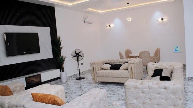 A modern living room with a flat-screen TV, comfortable sofas, a dining table, and a fan. - DEOSEA APARTMENTS (Abuja)