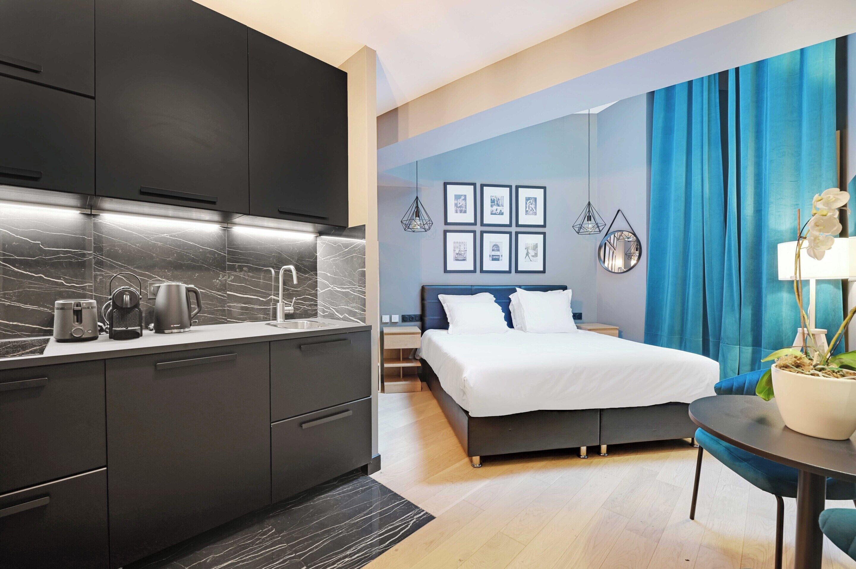 A modern hotel room with a kitchen area, a bed with white bedding, and a dining table with a potted plant.