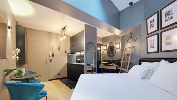 Quarto de hotel moderno com uma cama grande, uma escrivaninha com cadeira, um pequeno espaço de refeições e uma cozinha ao fundo.