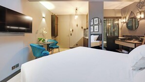 Quarto de hotel moderno com uma cama grande, televisão na parede, um pequeno espaço de estar com uma cadeira e uma mesa, e banheiro com espelho e iluminação.
