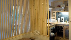 A room with a bed, a wooden slat partition, a small table with a vase, and a kitchenette area with a microwave and sink.