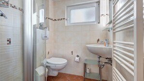 Een badkamer met een douchecabine van glas, een toilet, een wastafel en een raam boven de wastafel.