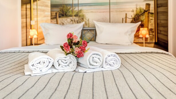 Een opgemaakt bed met opgerolde witte handdoeken en een vaas met roze bloemen.