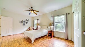 Une chambre avec un plancher en bois, un lit, une table de chevet, une chaise, un ventilateur de plafond et une fenêtre avec des rideaux.