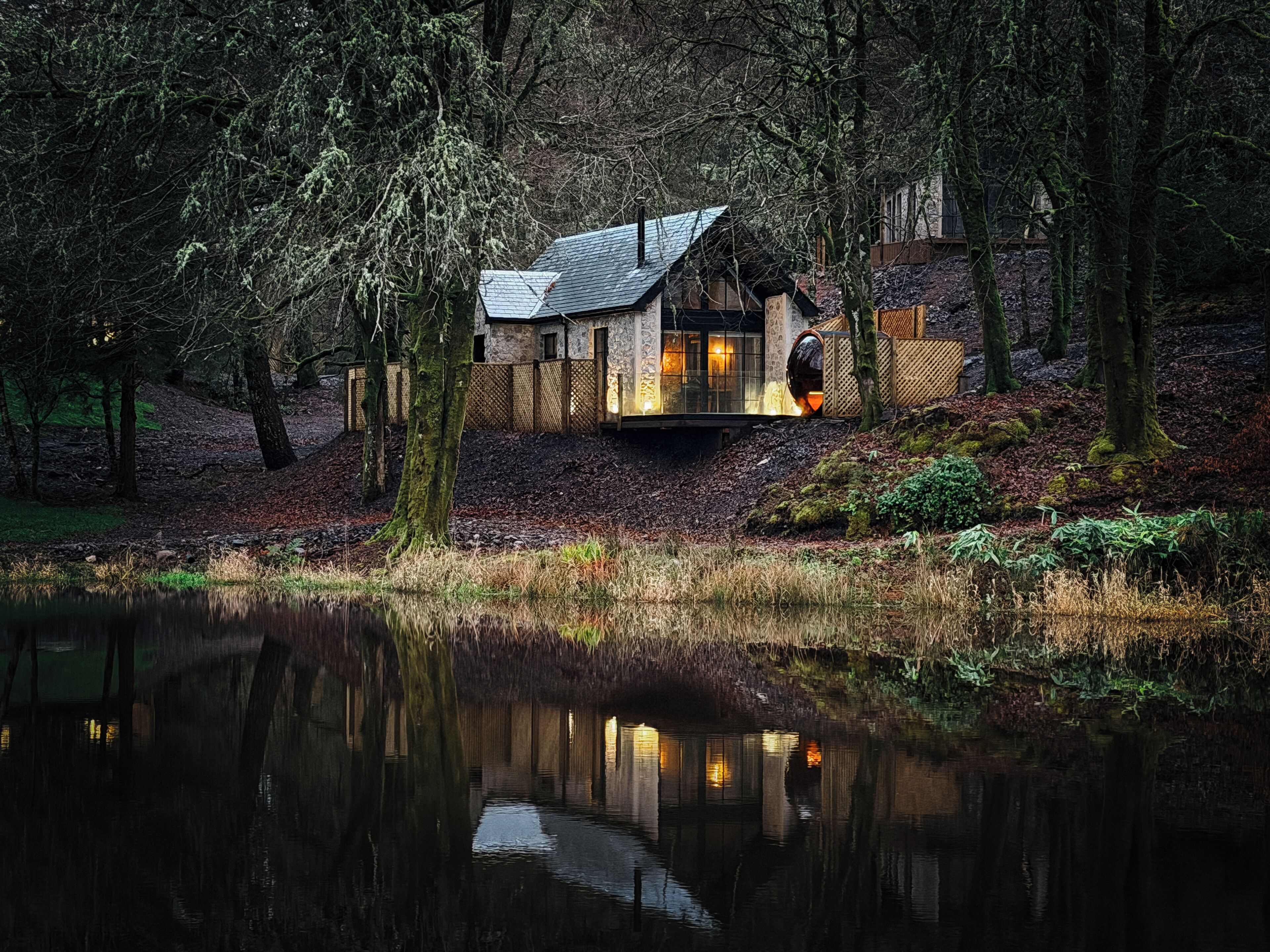 A rustic cabin with a stone exterior, a wooden deck, and a pond in the foreground.