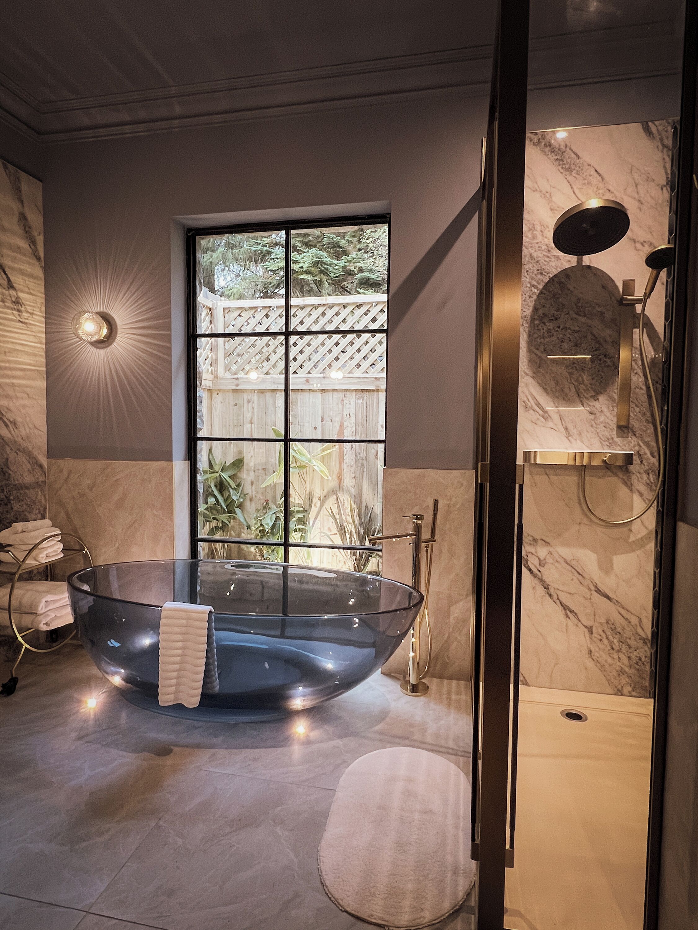 A modern bathroom with a glass bathtub, a large window with a view of greenery, and a marble wall.