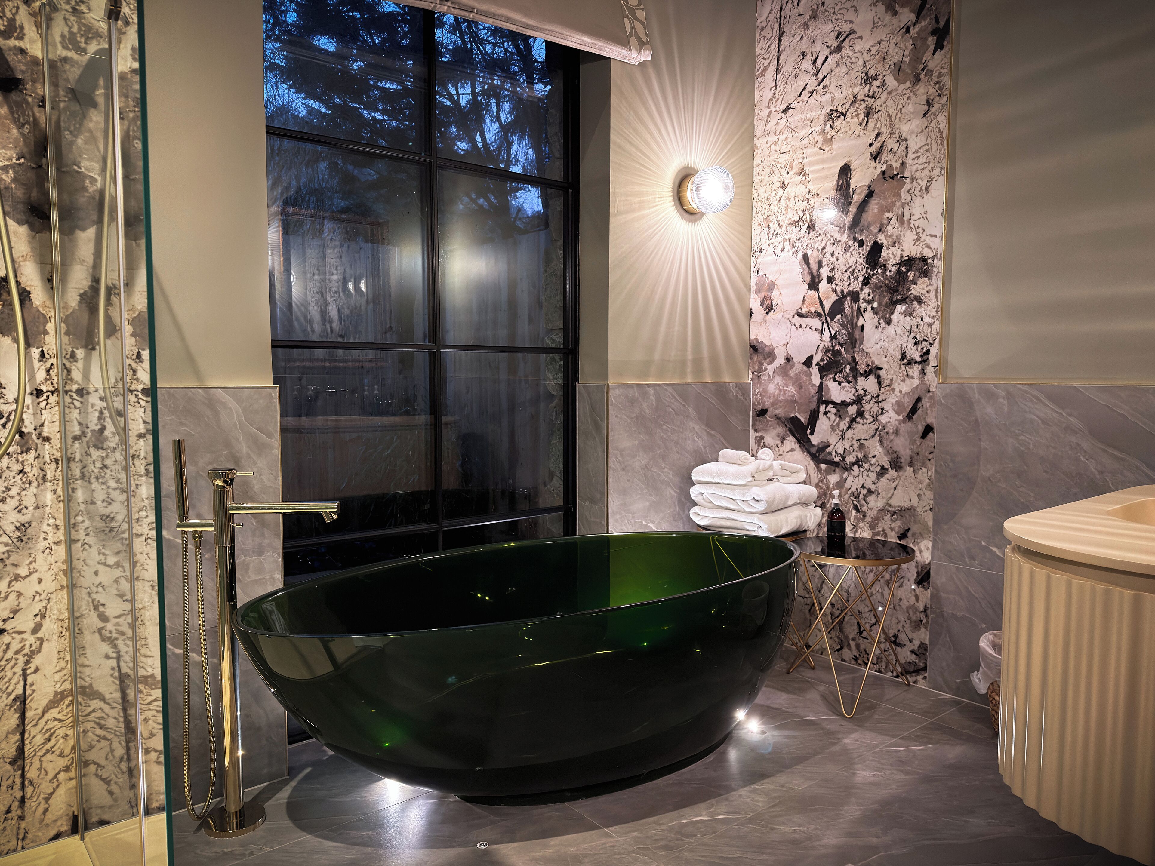 A modern bathroom with a large green bathtub, a glass shower enclosure, and a marble wall with a unique pattern.