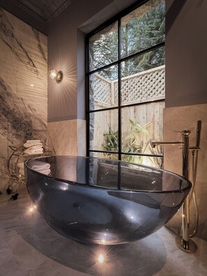 A modern bathroom with a large, clear bathtub, a wooden lattice window, and a marble wall. - Glencruitten House (Oban)