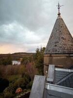 A building with a conical roof and a weather vane, surrounded by a landscape with trees and a lake.