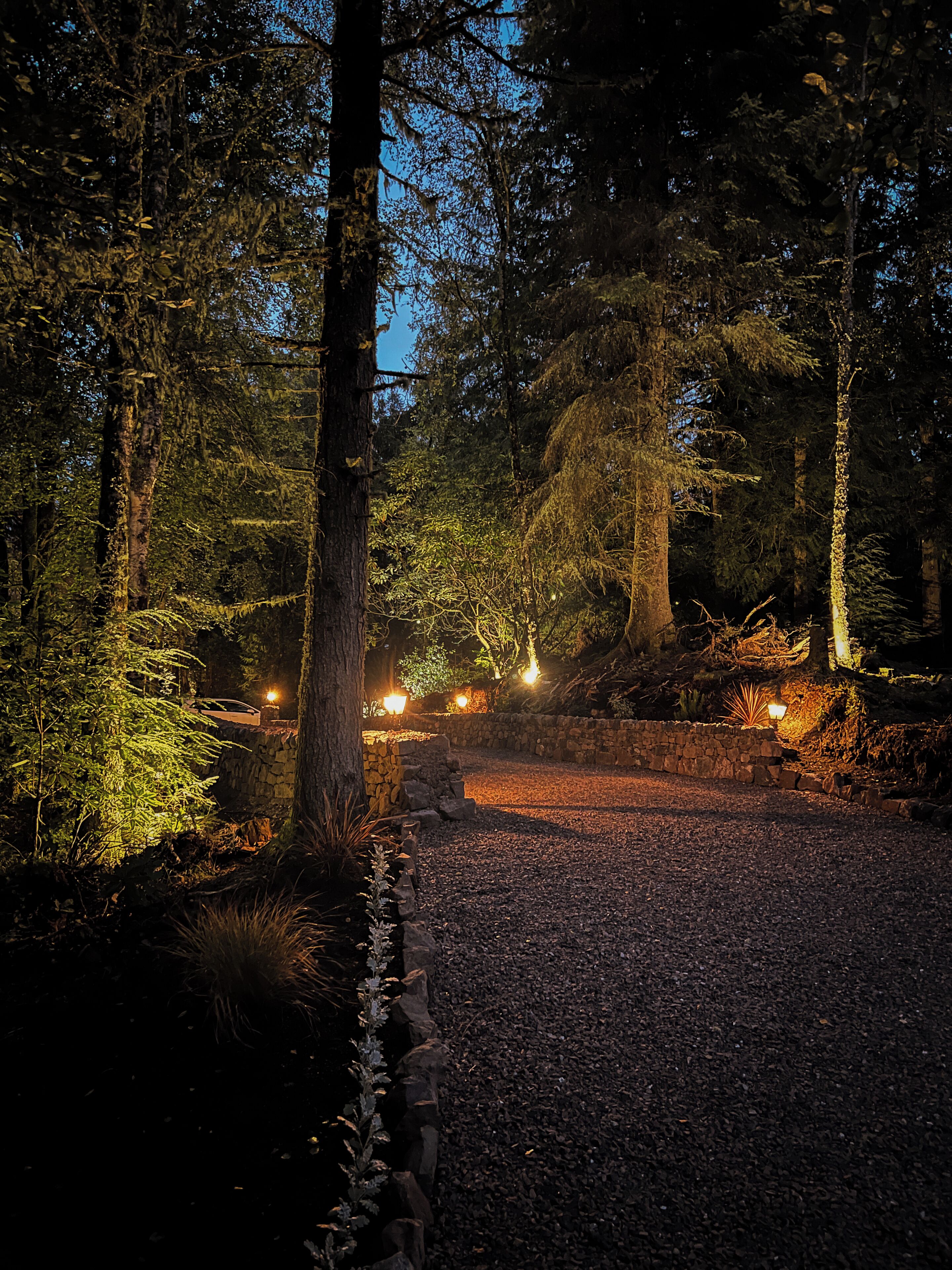 Ein bewaldetes Gebiet mit einem Schotterweg, Steinmauern und beleuchteten Lichtern.