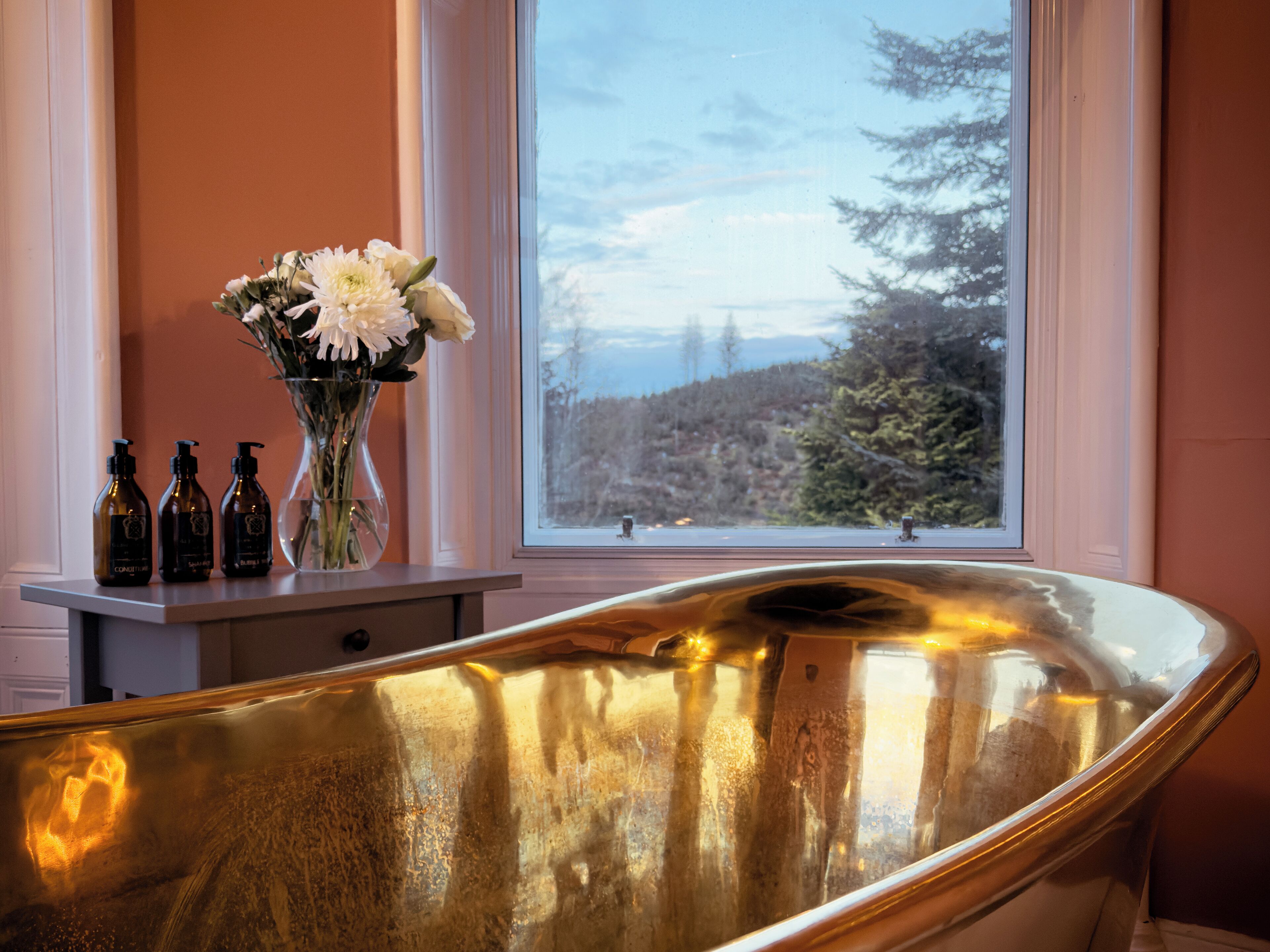 A bathroom with a freestanding bathtub, a vase with flowers, and three bottles on a shelf.