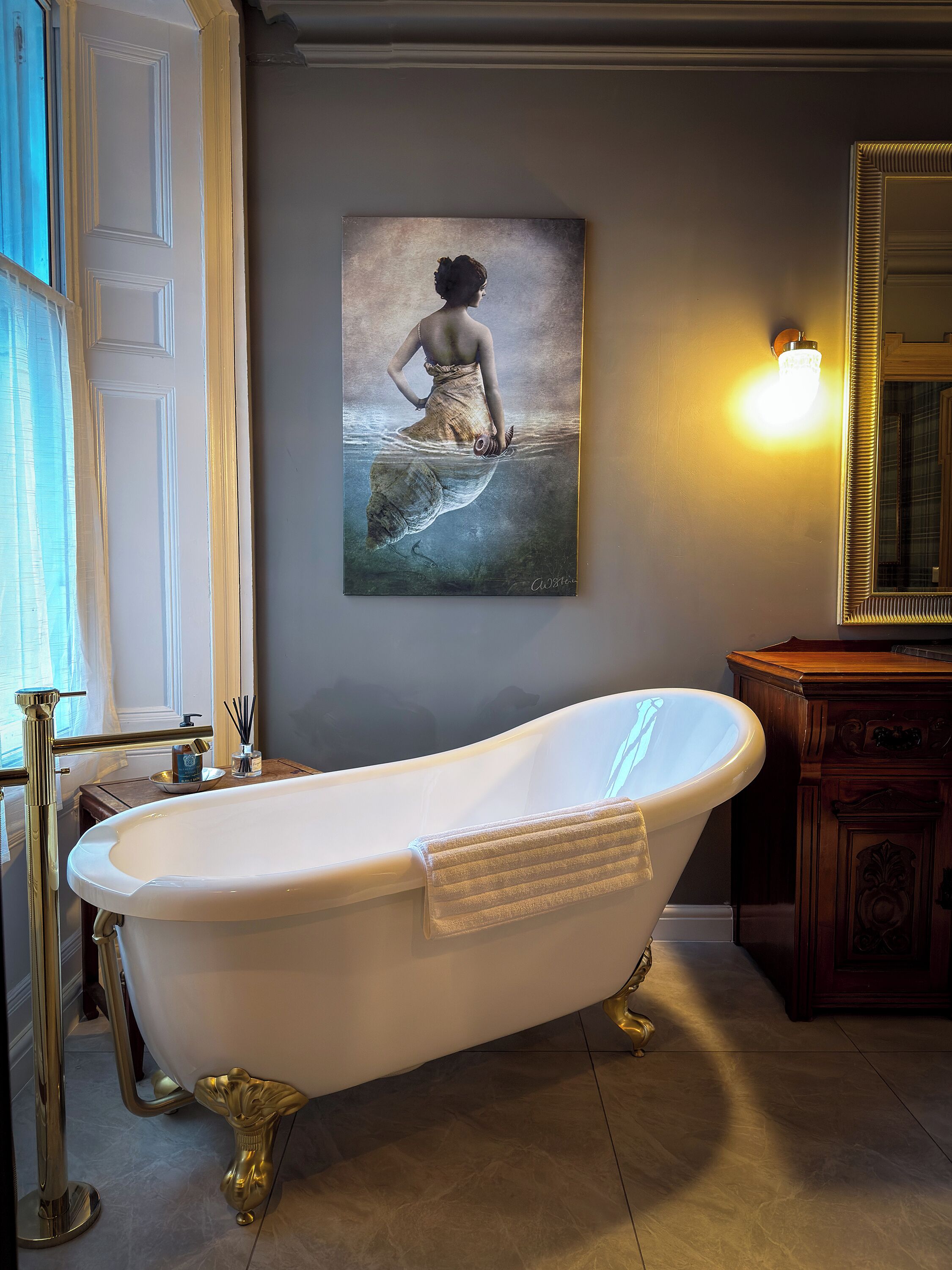 A bathroom with a freestanding bathtub, a large mirror, and a framed artwork on the wall.