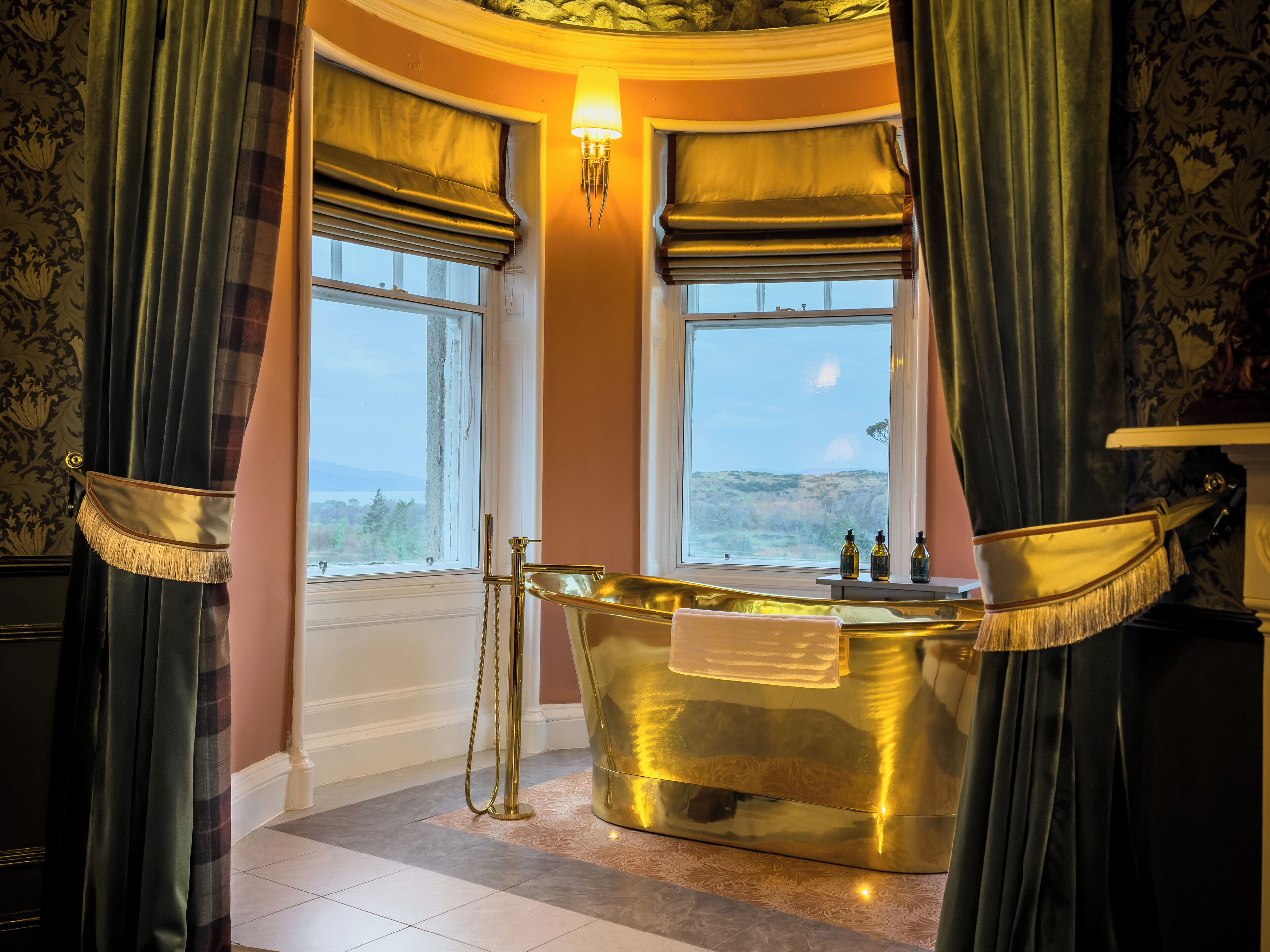 A luxurious bathroom with a gold bathtub, two windows with views, and ornate curtains.