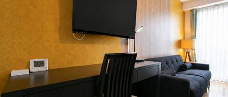 A modern hotel room with a flat-screen TV mounted on the wall, a desk with a chair, a sofa, and a lamp.