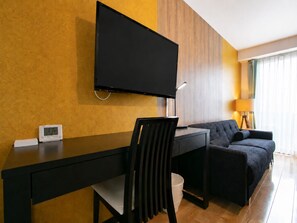Ein modernes Hotelzimmer mit Flachbildfernseher an der Wand, einem Schreibtisch mit Stuhl, einer Couch und einer Lampe.