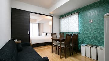A compact hotel room with a bed, a dark-colored sofa, a dining table with chairs, and a small kitchenette.