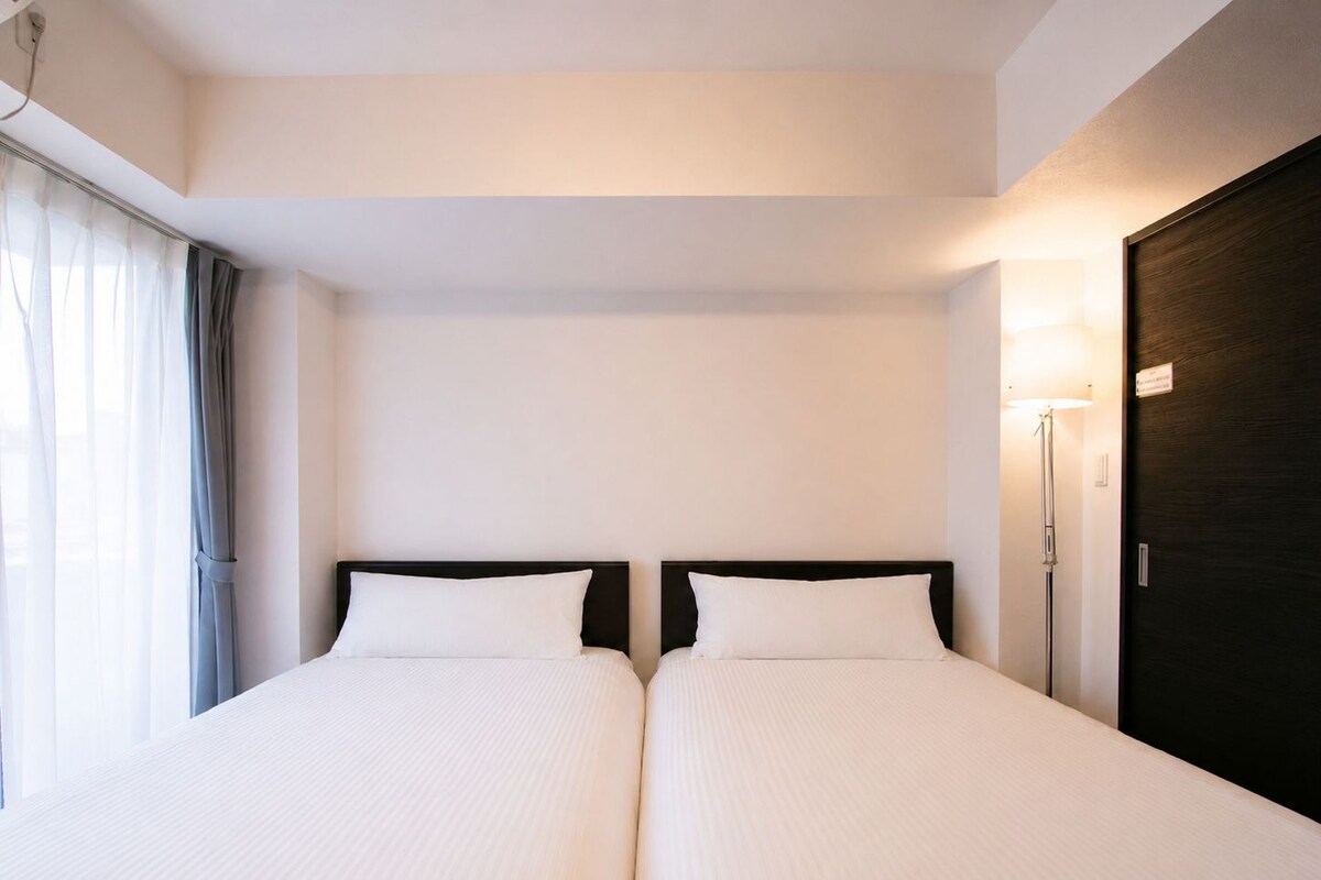 Une chambre d’hôtel avec deux lits, une fenêtre avec des rideaux, une lampe de chevet et une porte de couleur sombre.
