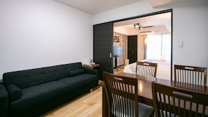A modern living room with a dark sofa, wooden dining table, and chairs. A TV is mounted on the wall, and there is a door leading to another room. - Randor Residence Tokyo Annex (Tokyo)
