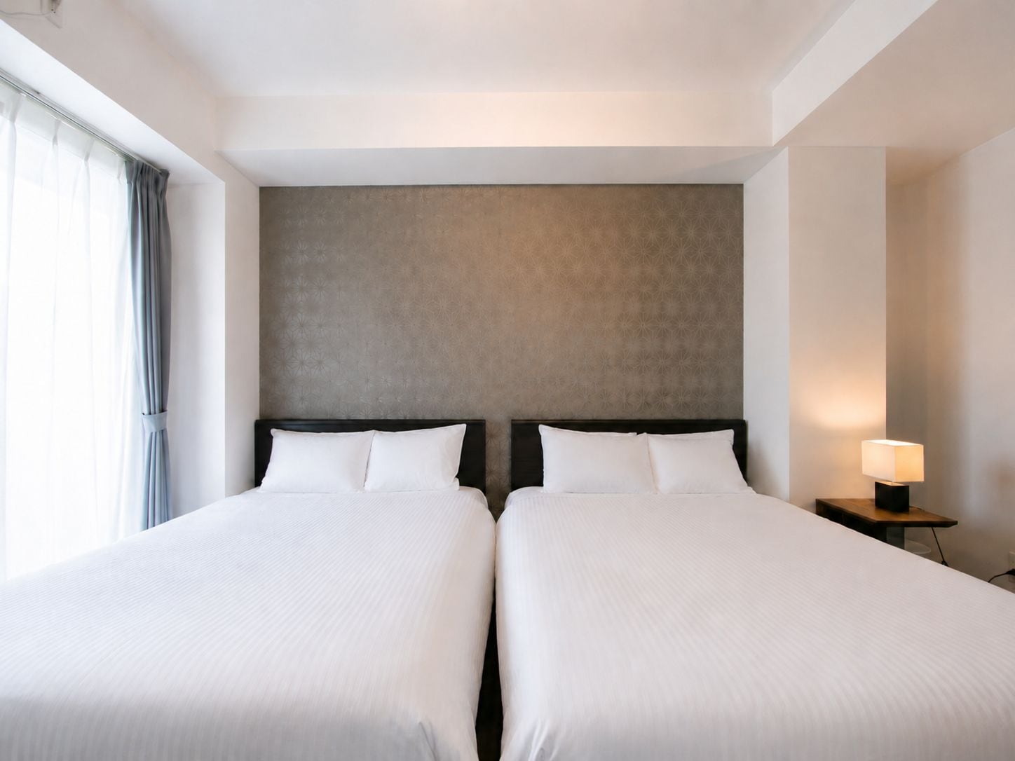 A hotel room with two beds, a nightstand with a lamp, and a large window with curtains.