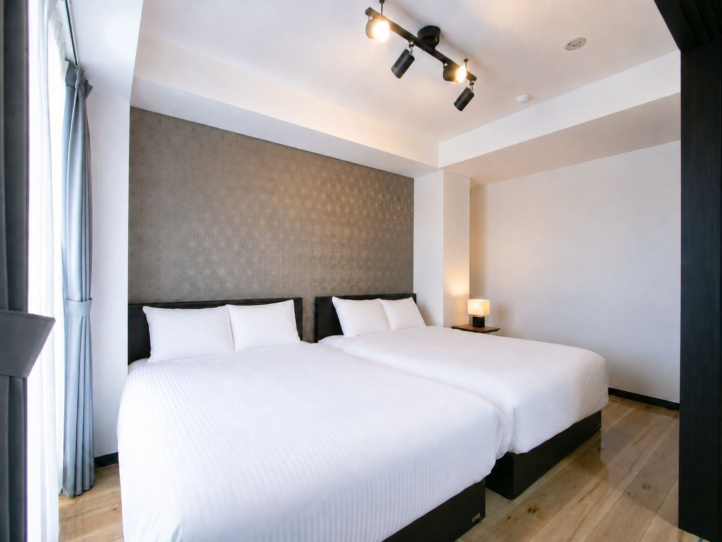 A hotel room with two beds, a patterned wall, a ceiling light fixture, and a bedside table with a lamp.