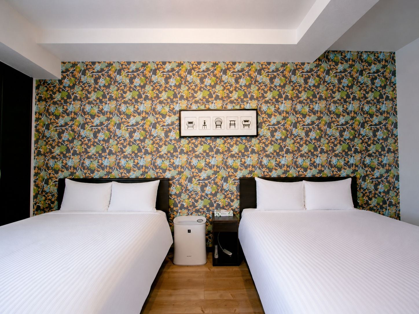 Habitación de hotel con dos camas, una mesita de noche, un cubo de basura y una pared con estampado floral.