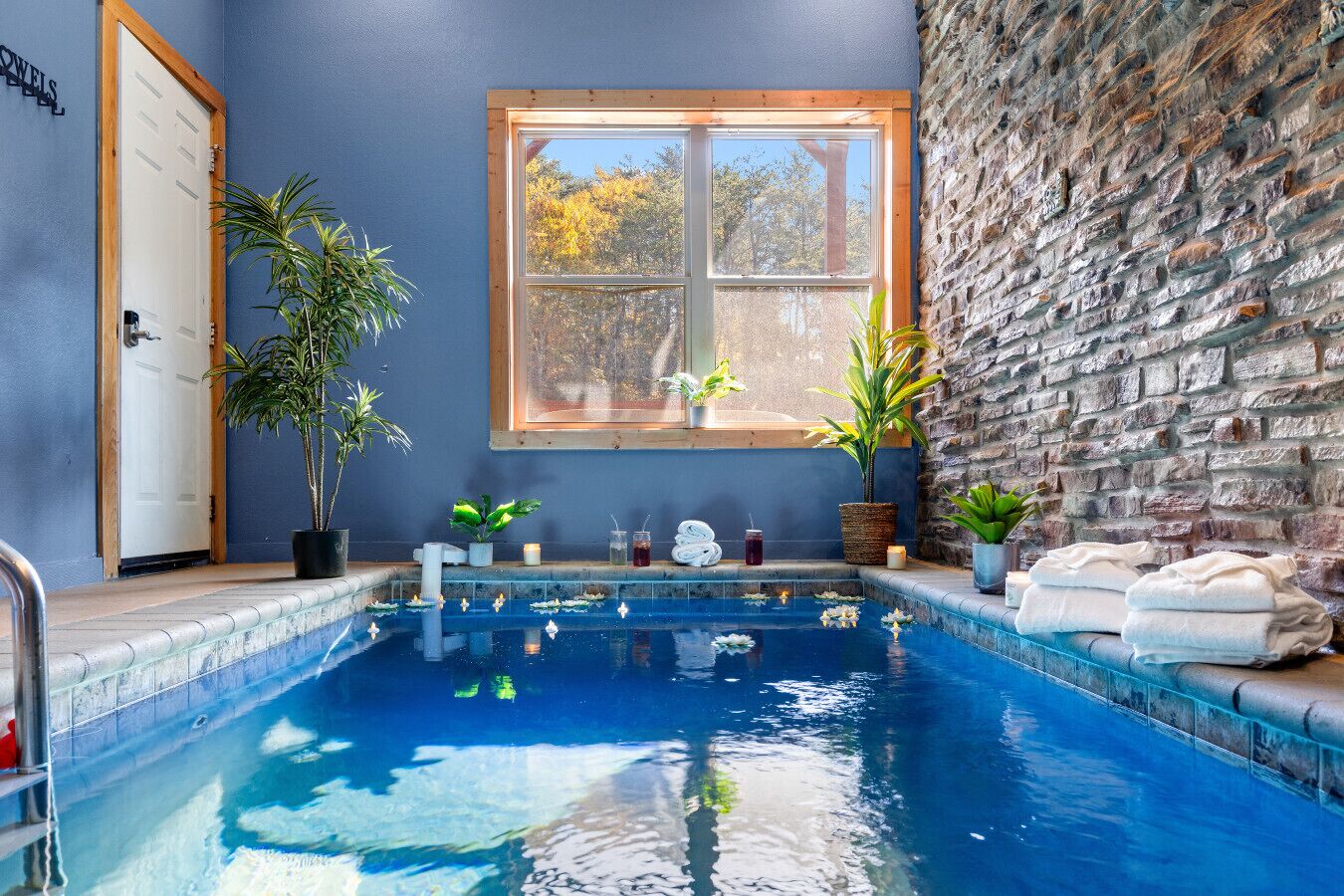 Une piscine intérieure avec un mur en pierre, une fenêtre donnant sur les arbres, ainsi que des serviettes et des plantes disposées au bord du bassin.