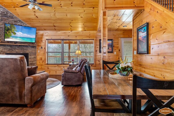 A wooden cabin interior with a stone fireplace, a TV mounted on the wall, and a dining area with a table and chairs.