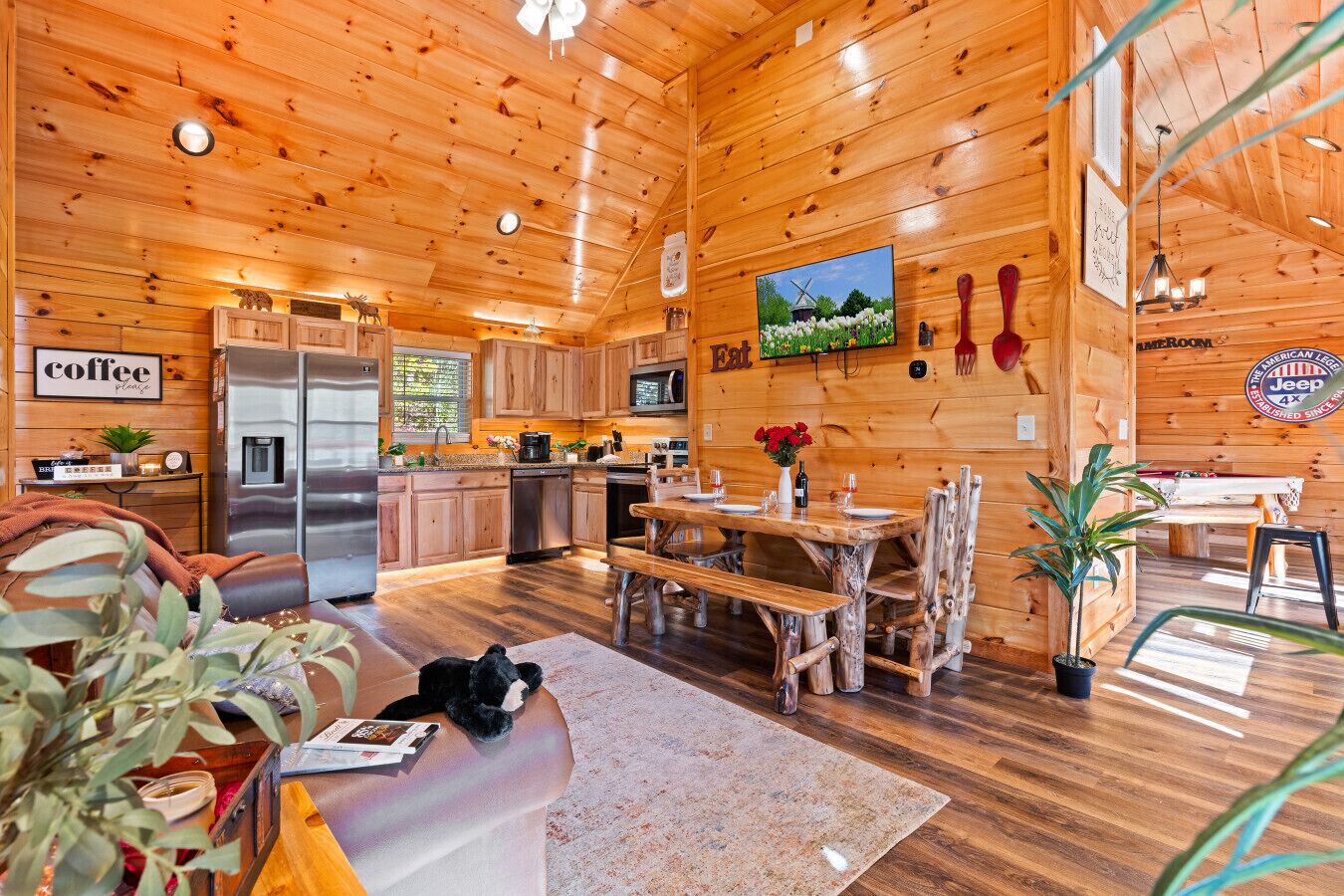 A cozy cabin interior with wooden walls, a kitchen area, a dining table, and a living room with a sofa and a dog.