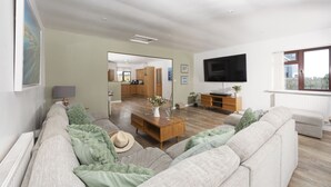 A modern living room with a sofa, coffee table, and TV. There is a kitchen in the background and a large window. - Wheal Frances -Hot Tub - Free Parking - Free Wi-Fi - Accessible (Helston)