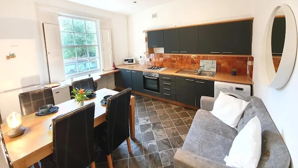 A modern kitchen with dark cabinets, a dining area with a table and chairs, and a large window providing natural light. - House in Central London - Sleeps up to 16 (London)