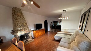 A living room with a stone fireplace, a sofa, a TV, and a kitchen area in the background. - Ideal Holiday Rental House for Families Near the Beach (Calonge)