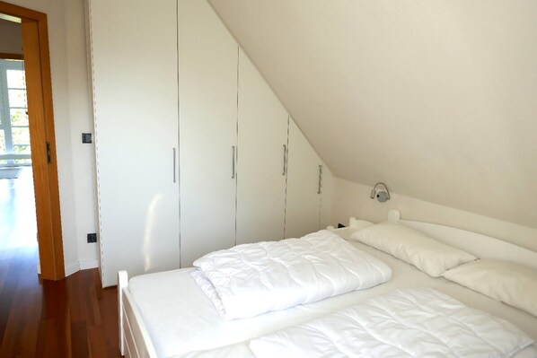 A bedroom with a sloped ceiling, white bedding, and a wardrobe with silver handles.
