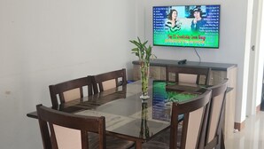 Un comedor con una mesa de vidrio rodeada de sillas de madera, una planta pequeña en maceta y un televisor mostrando una lista de reproducción musical.