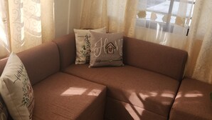 Un sofá seccional marrón con almohadones decorativos, ubicado frente a una ventana con cortinas traslúcidas.