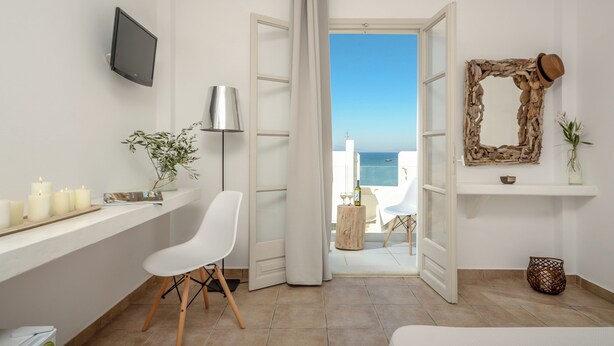 Une pièce avec une télévision, une chaise blanche, une petite table avec des bougies et une vue sur la mer par une porte ouverte.