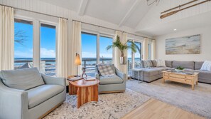 A spacious living room with a large window overlooking the beach, a sofa set, a coffee table, and a painting on the wall.
