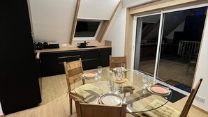 A modern dining area with a glass table, wooden chairs, and a kitchen in the background. - Guergonet, Cosy 2-bedroom accomodation (Châtelaudren-Plouagat)