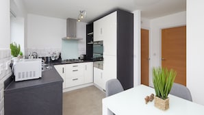 Eine moderne Küche mit weißen Schränken, einer schwarzen Arbeitsplatte und einem eingebauten Backofen. Eine kleine Kücheninsel beherbergt eine Kaffeemaschine und einen Blumentopf.