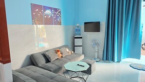 A modern living room with a grey sofa, a small round table, a water dispenser, a standing fan, and a TV mounted on the wall.