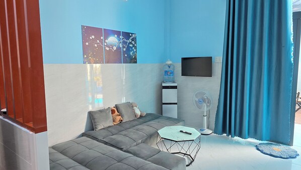 Una sala de estar moderna con un sofá gris, una mesita redonda pequeña, un dispensador de agua, un televisor montado en la pared y un ventanal con cortinas azules.