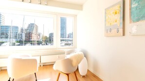 A modern living room with a large window, white furniture, and artwork on the wall. - Apartment With a View of the Marina (Ostend)