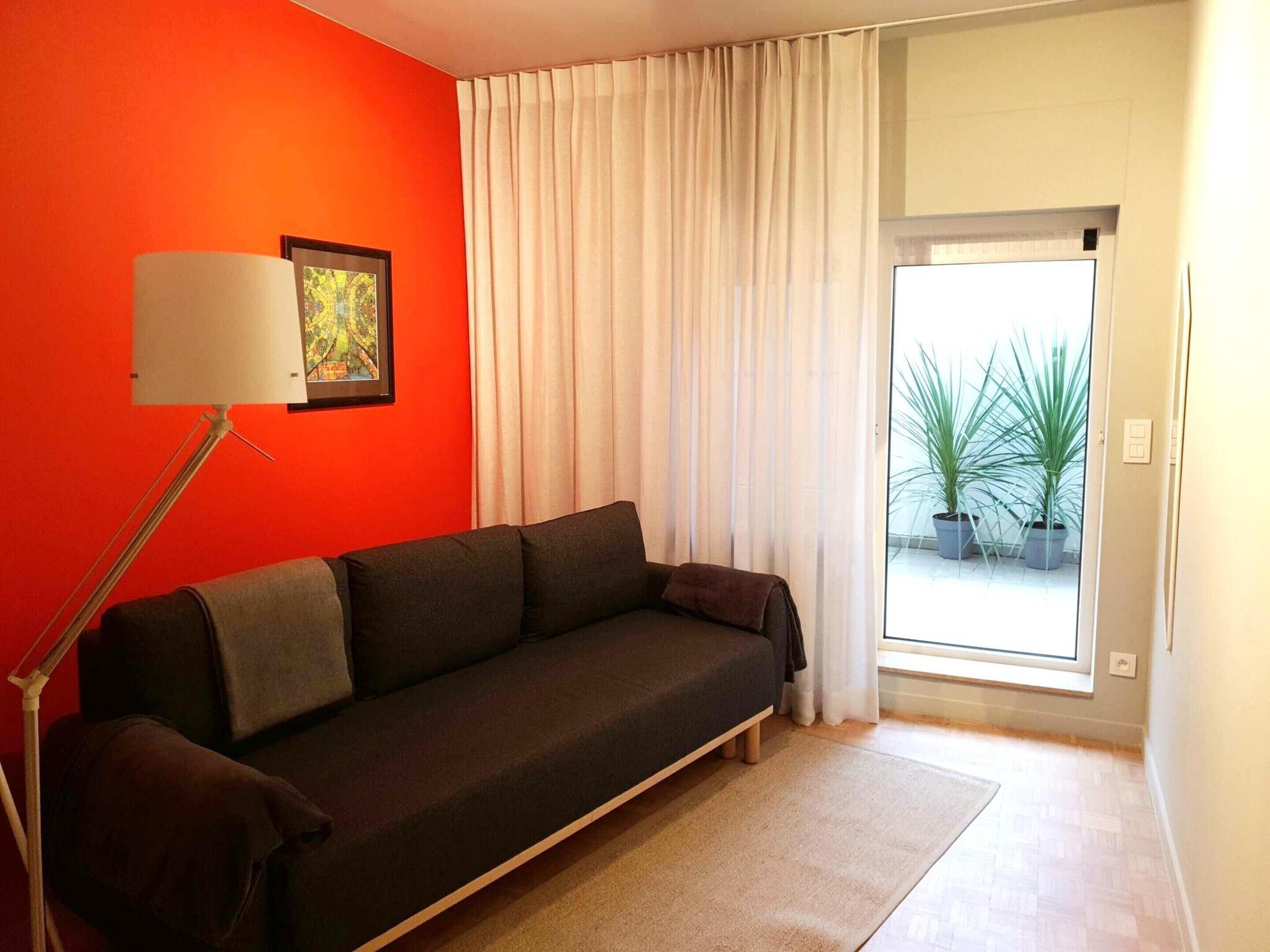 A modern living room with a dark sofa, a floor lamp, and a large window with sheer curtains.