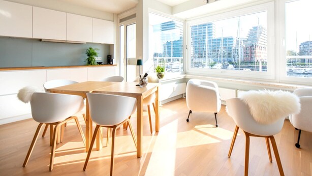 A modern kitchen with white cabinets, a wooden dining table, and chairs. There is a large window with a view of buildings and a marina. - Apartment With a View of the Marina (Ostend)