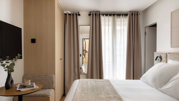 Una habitación de hotel con una cama, una mesita con un florero y una revista, una silla y un televisor en la pared.